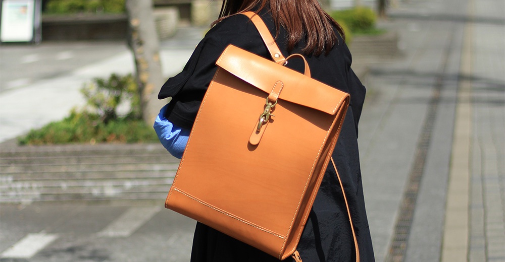 光滑皮革製成的棕色方形後背包，配有金屬扣環和可調節肩帶，由穿著黑色外套的人背著，背景為戶外街道。