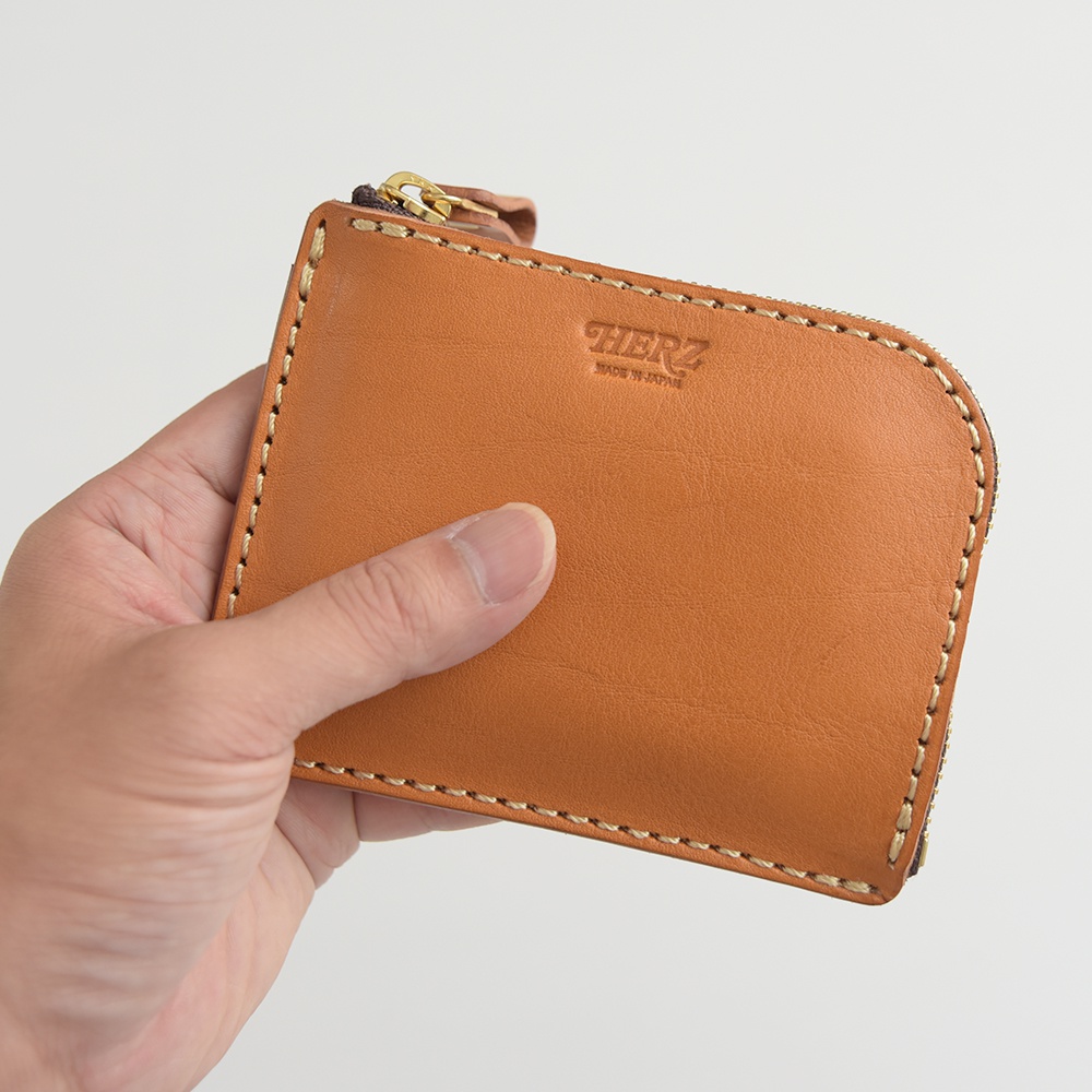 手持一個棕色皮革L型拉鍊迷你短夾，皮革表面有燙印品牌Logo。