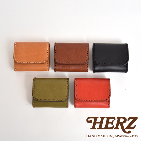 五個不同顏色的日本HERZ職人手作皮革零錢包整齊排列，呈現出棕色、咖啡色、黑色、綠色與紅色五種款式。每個零錢包皆採用雙折設計搭配對比色縫線，右下角印有HERZ品牌商標與日本手工製作年份字樣，整體展現質感細膩的皮革工藝風格。
