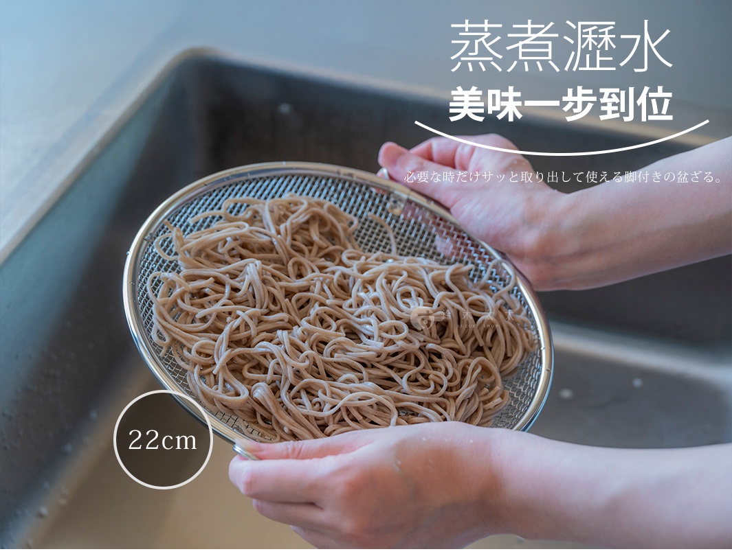 一雙手正捧著一個直徑22公分的不鏽鋼圓形瀝水篩網，網內裝有剛煮好的蕎麥麵，上方標註有蒸煮瀝水美味一步到位以及必要時隨手取用的帶腳瀝水籃等日文字樣，整體呈現於廚房流理台的背景中。