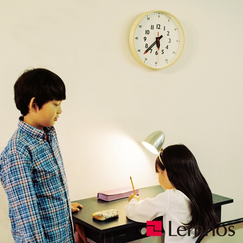 一個男孩穿著藍色格子襯衫，站在桌子旁，桌上有一盞檯燈、一個紫色筆盒、一個小化妝包和一個女孩在寫字。牆上掛著一個圓形白色蒙特梭利時鐘，帶有黑色數字和紅色秒針。