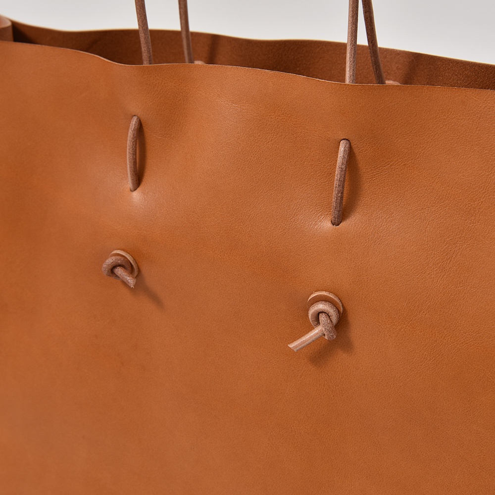 棕褐色皮革提袋，有細緻的皮繩穿孔與打結設計，皮革材質柔軟，展現職人手作質感。