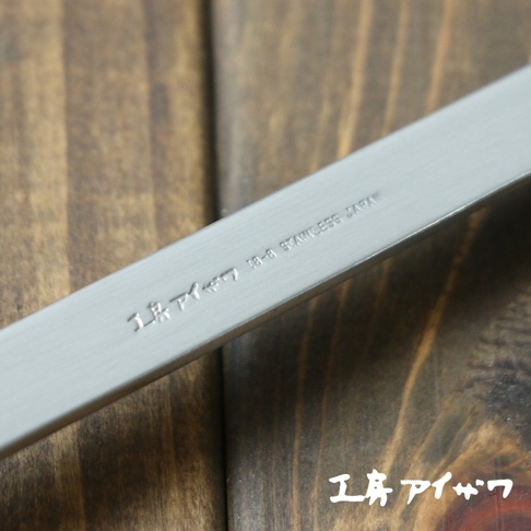 不鏽鋼萬用鍋鏟特寫，銀色金屬材質，鍋鏟上蝕刻有「18-8 STAINLESS JAPAN」字樣及日文品牌標示，置於深色木紋背景上。