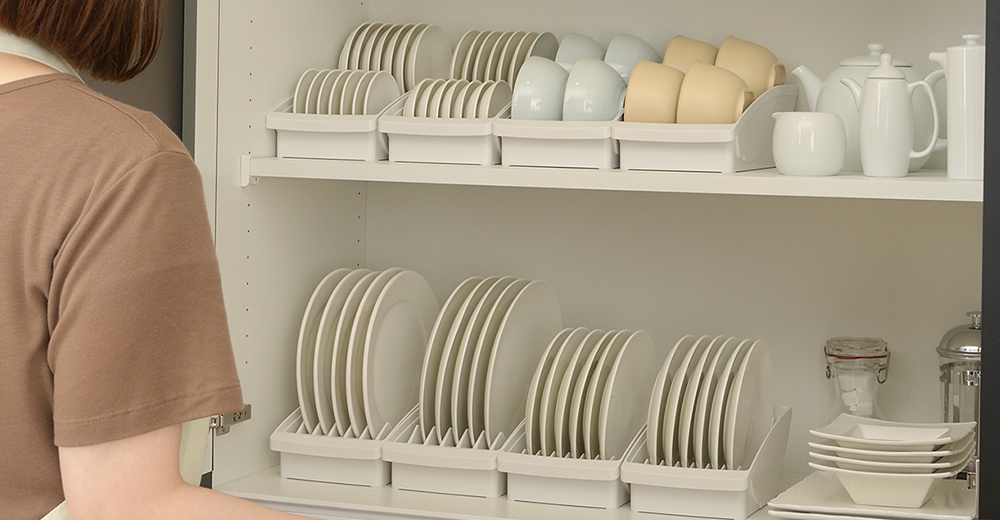 櫥櫃裡整齊擺放著白色餐盤架，架上收納著各式大小的盤子、杯子及茶壺組，畫面右側有一名身穿咖啡色上衣的女子。