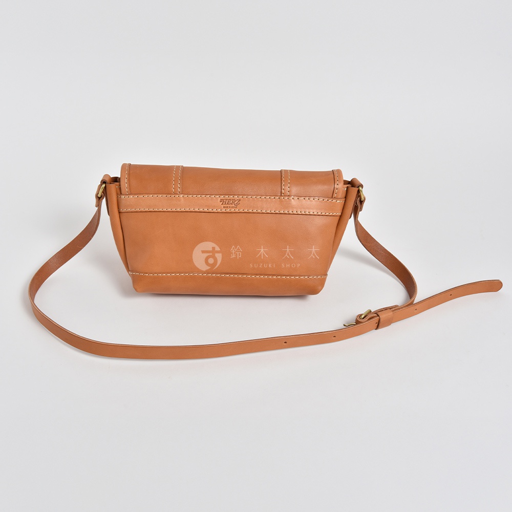 職人手作皮革復古郵差包，採用棕褐色牛皮製成，包身結構紮實，邊緣有明顯的縫線，搭配可調節長度的皮革背帶，包包側面可見品牌標誌。