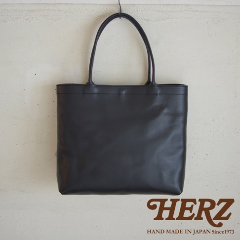 黑色皮革質感的托特包，簡約設計，有兩個提把，包身線條俐落，下方有品牌標誌HERZ。