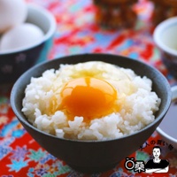 一碗白米飯上放著一顆生雞蛋黃，雞蛋黃呈亮橘色，周圍點綴著一些蛋白。背景是紅底、印有彩色花紋的布料。