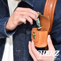 職人手作皮革耳機收納包，淺棕色，附黃銅釦，展示將潤唇膏放入包內的動作。