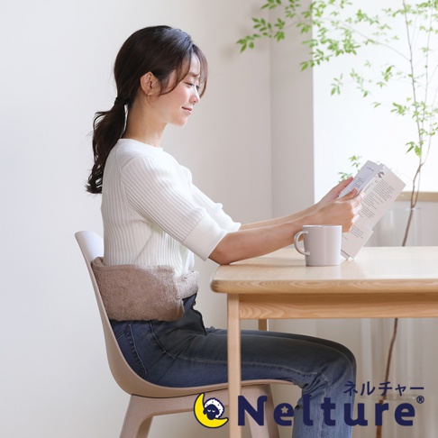 一名女子坐在椅子上，腰部圍著一個淺棕色的毛絨腰枕，她正閱讀手中的書本，桌上放著一杯咖啡。