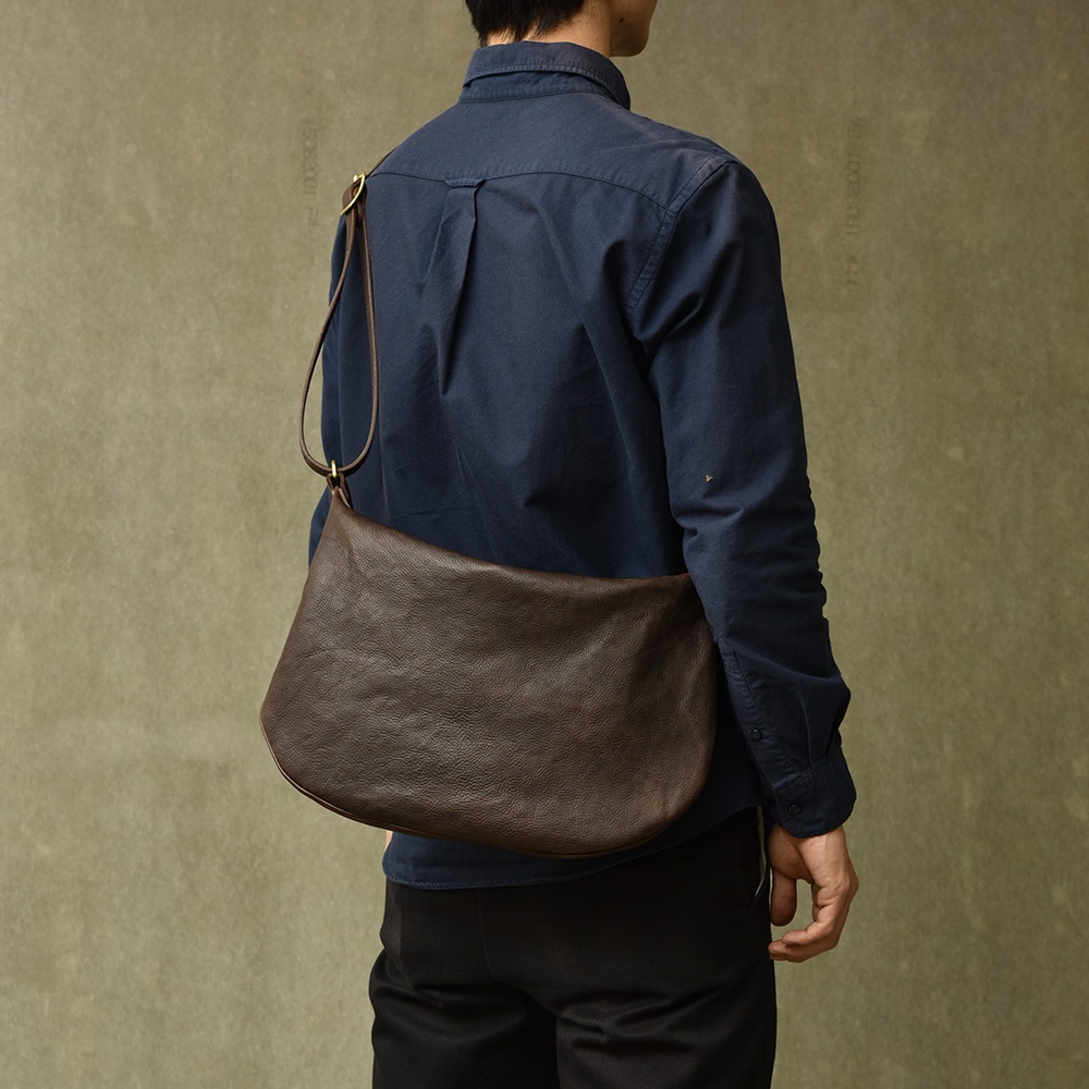 職人手作深棕色皮革單肩半月包，斜背於身穿海軍藍襯衫的模特兒身上，包款線條圓潤，皮革表面有自然紋理。