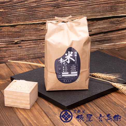 一個棕色紙袋裝著北海道七星米2公斤，袋頂繫著麻繩。袋上有深藍色標籤，標示著「現職米」和「幸」字樣，以及其他產品資訊。一旁有一個木盒裝著白米，背景是木紋和麥穗。