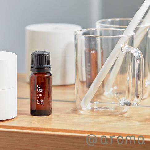 一個深褐色玻璃瓶裝有 C03 純天然木香精油，瓶身標有白色字體「C 03 Clean air Clean FOREST」。瓶蓋為黑色。背景是白色圓柱形物體和透明玻璃杯。