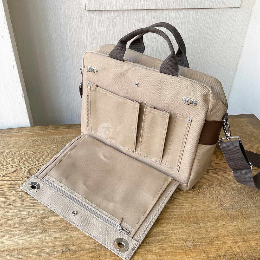 犬印純綿帆布三用橫式側背包，米白色帆布材質，搭配深棕色皮革飾邊與提把，設有多個外袋與可拆卸肩帶，另有可攤開的內層設計，包含拉鍊口袋。