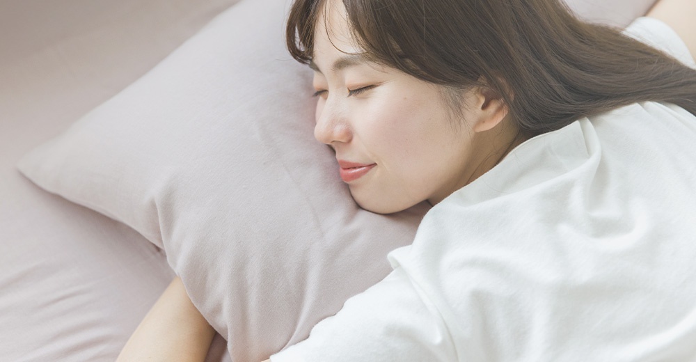 一位年輕女性穿著白色上衣，閉著眼睛安穩地躺在淡粉色的枕頭上，呈現出寧靜且放鬆的睡眠畫面。