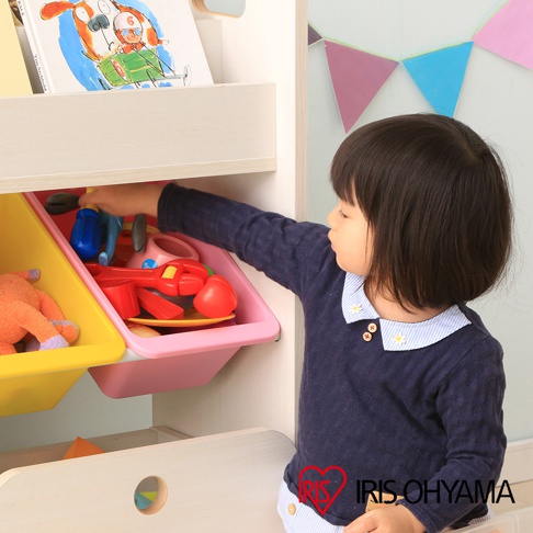 木製童心玩具繪本收納架，附有黃色與粉紅色兩個塑膠收納盒，盒內裝有玩具。左側收納盒內有橘色絨毛玩具，右側收納盒內有紅色扳手、藍色水管等玩具。一名幼童正伸手從粉紅色收納盒拿取玩具。