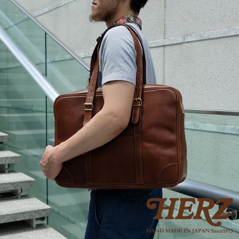 一位男士肩背一個棕色皮革製成的兩用公事包，包包有手提把和肩背帶，側邊有拉鍊，整體呈現質樸的風格。