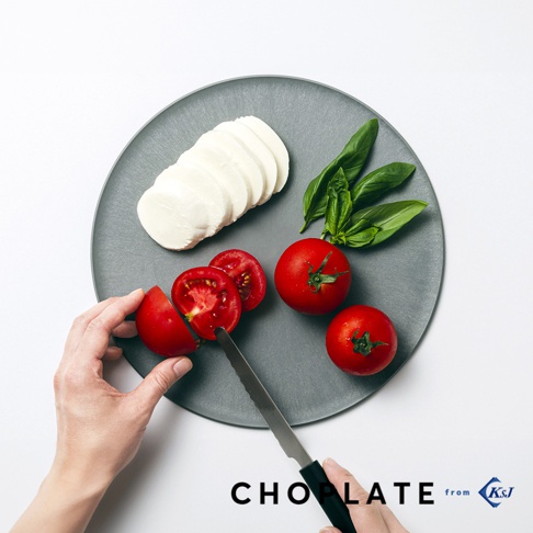 灰色圓形兩用砧板盤上，擺放著切片莫札瑞拉起司、聖女番茄、羅勒葉，以及正在被刀切開的番茄。