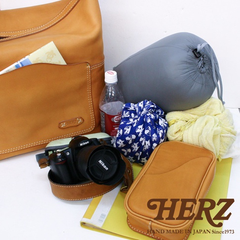 鞣革厚實單肩波士頓包，搭配一個棕色皮革側袋、一個藍白貓咪圖案束口袋、一個灰色壓縮袋、一瓶飲用水、一條黃色絲巾及一台黑色 Nikon 相機。