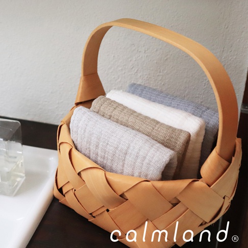 編織木質提籃裡整齊疊放著四條長方巾，顏色從左至右分別為淺灰、米駝、純白、淺藍。布料表面帶有細緻的紋理，展現無撚紗的質感。