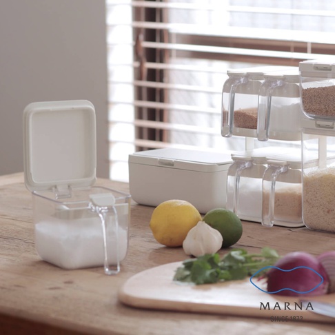 木質桌面擺放著 Marna 品牌的透明密封調味料盒，盒內裝有白色粉末，蓋子向上開啟。旁邊還有一個長方形白色收納盒，以及一組堆疊的透明儲物罐，罐中盛裝各種調味料。前景放置了檸檬、青檸、大蒜、香菜與紅洋蔥，畫面整體呈現清新簡約的廚房收納風格。