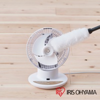 白色 PCF-SC15T 空氣循環扇，擁有簡約的圓形底座和傾斜角度可調的白色扇網，風扇中心有一個白色扇葉，一個白色毛刷正伸入扇網清潔。產品放置在淺木紋地板上，右下方有 IRIS OHYAMA 的紅色心形 Logo。
