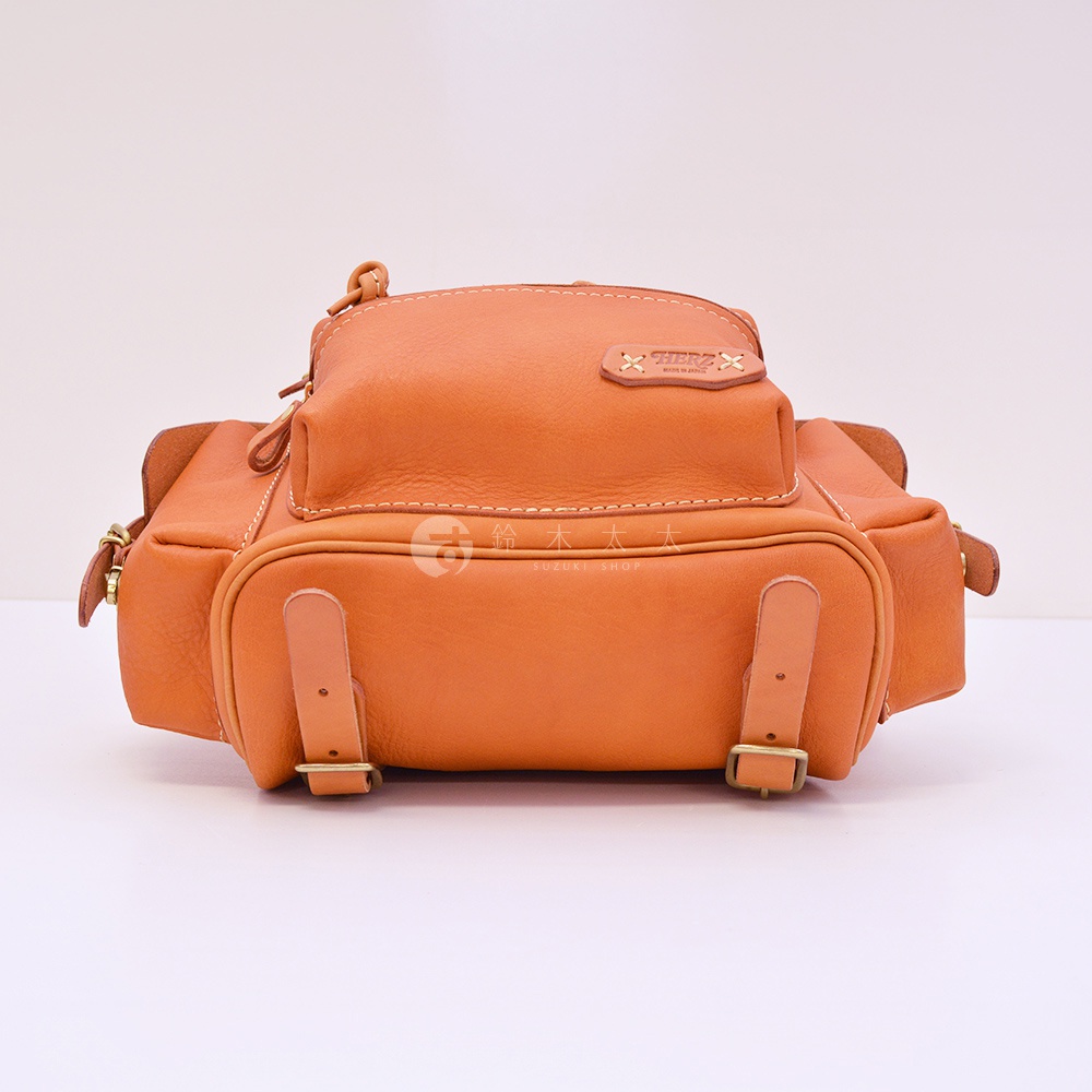 職人手作皮革復古風旅行後背包，呈現溫暖的橘棕色調，包身採用天然皮革，帶有明顯紋理與縫線細節，外觀設計簡潔實用，配有金屬扣環和皮革束帶。