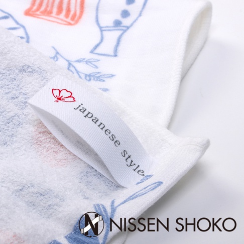 白色棉紗廚房拭巾，印有日式風格的藍色與橘色花卉及植物圖案。一條白色織帶標籤縫於拭巾上，印有紅色小花和「japanese style」字樣。