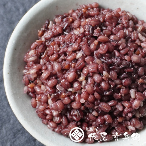 一碗裝滿煮熟的日本國產十六穀米，色澤呈現天然的紫紅色。米飯顆粒飽滿且質地豐富，混合了多種穀物，表面帶有微微的光澤，視覺上展現出Q彈的口感，右下角印有三代目俵屋玄兵衛的品牌標誌。