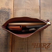 職人手作皮革Organ拉鍊式小物收納袋，展示棕色皮革材質，袋內裝有卡夾、筆記本、手機及鑰匙等小物，拉鍊為金色，整體置於木紋背景上。