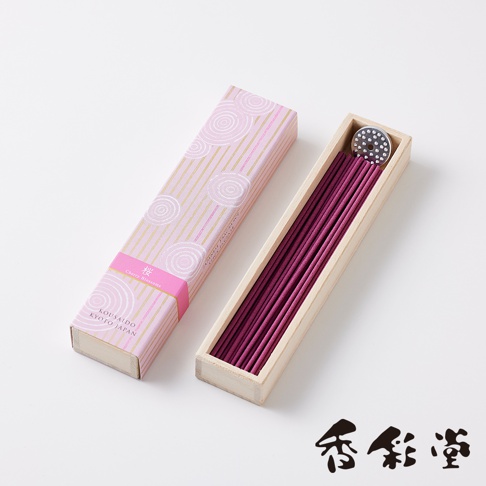 粉色包裝盒內裝有數支紫紅色線香，盒蓋上印有金色線條和粉色螺旋圖案，下方標有「KOUSAIDO KYOTO JAPAN」字樣。線香置於淺木色長方形木盒中，盒內有一個帶有銀色圓點的圓形金屬蓋。