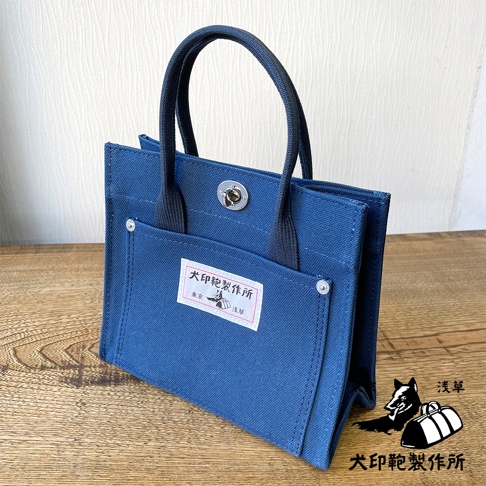 藍色純棉帆布迷你托特包，設有兩個提把、正面有兩個外袋，其中一個外袋上貼有印有日文文字和圖案的標籤，包包開口處有金屬旋轉鎖扣。