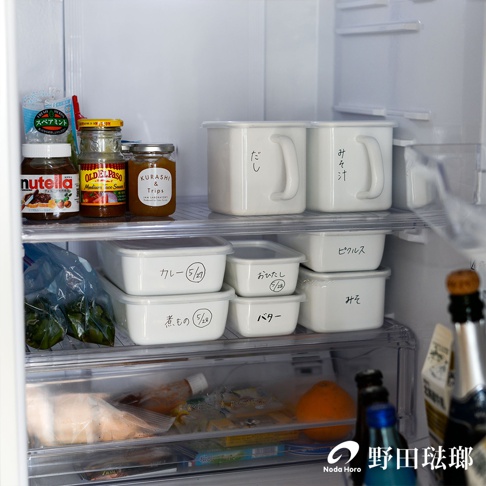 冰箱內擺放著多個長方型淺型白色琺瑯保存容器，其中兩個較大的容器帶有把手，分別寫有「だし」和「みそ汁」的日文。下方另有幾個較小的白色保存容器，標示著「カレー」、「おひたし」、「煮もの」、「バター」和「みそ」等內容物名稱及日期。