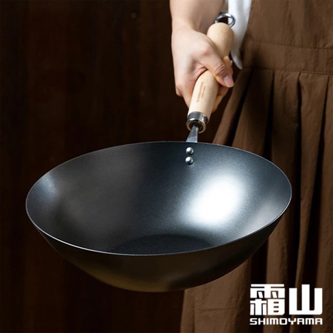 一只手正拿著日本製33公分木柄窒化鐵炒鍋，鍋體為黑色，鍋柄為原木色，鍋身有兩顆鉚釘。