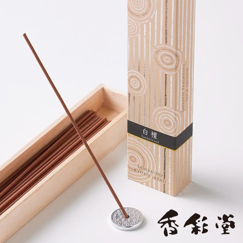 京都百樂香白檀香氛線香，淺木盒內裝有多支棕褐色線香，其中一支立於銀色圓形香座上。產品包裝盒為米白色，飾有銀色線條和螺旋紋，印有「白檀」及日文、英文產品資訊。