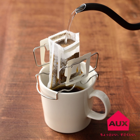 銀色金屬製可調式耳掛咖啡專用架，夾著濾掛式咖啡包，滴漏至白色馬克杯中，背景為木紋桌面。