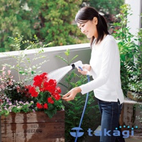 一位女子手持白色與藍色的灑水槍，正為木製花槽中的紅色花朵澆水，藍色水管連接灑水槍。