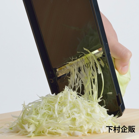 手持黑色超薄切高麗菜刨絲器，正在刨切新鮮高麗菜，刨出的細絲堆疊成一堆。