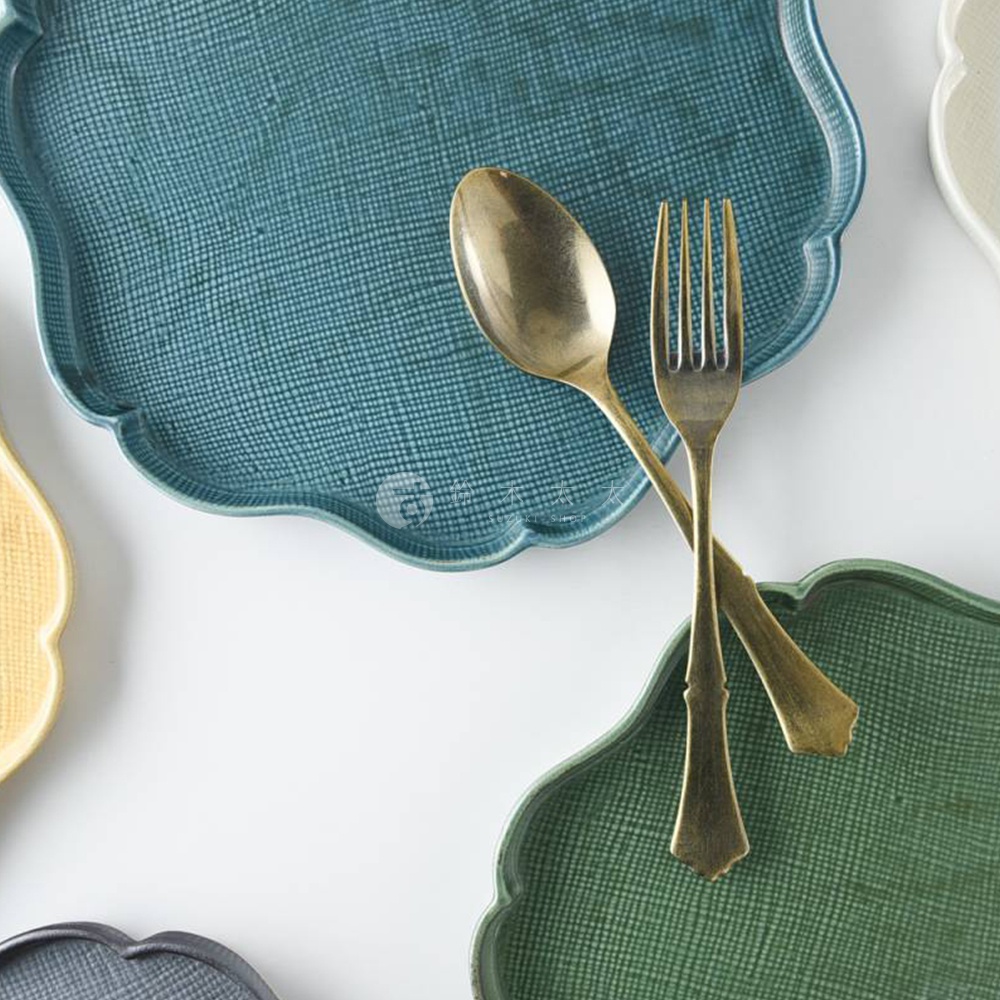 美濃燒花型主食盤，表面有細緻的網狀紋理，盤身邊緣呈花瓣狀。圖片中可見藍色、綠色與黃色等顏色的盤子，以及一組金色湯匙與叉子。