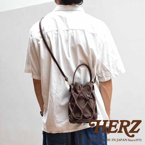 日本HERZ職人手作皮革Organ編織水桶包 G-98，模特身穿白色寬鬆襯衫，側背著棕色皮革與帆布拼接的編織水桶包，包包上有流線型的皮革裝飾，五金件閃亮，包包頂部有束繩開口。