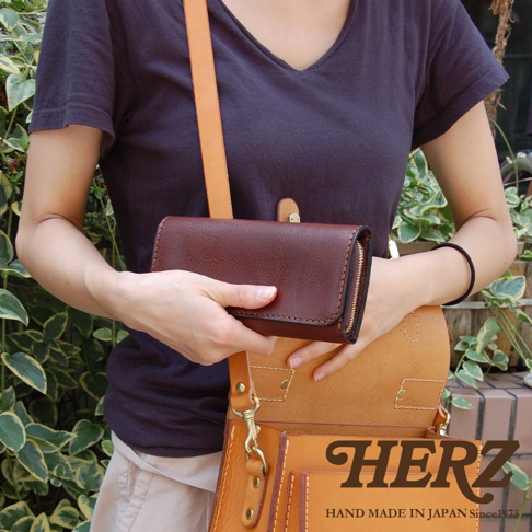 一位女性手持一個深棕色皮革長夾，夾子上有拉鍊。她身穿深藍色上衣，斜背一個淺棕色皮革側背包，包包上有「HERZ」和「HAND MADE IN JAPAN Since1973」的字樣。
