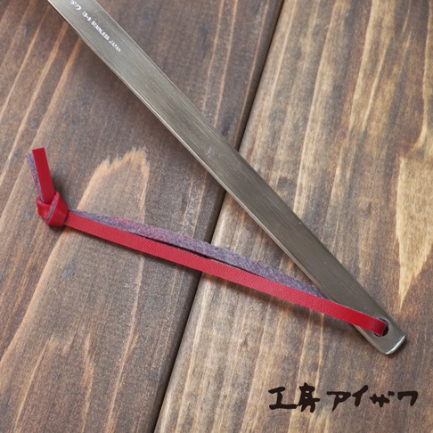 不鏽鋼萬用撈泡勺，勺柄為銀色霧面不鏽鋼材質，末端繫著紅色皮革與灰色絨布編織的掛繩。