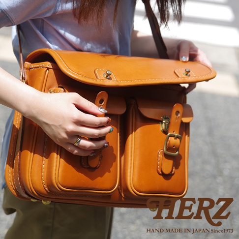 日本HERZ手工皮革雙扣郵差包，棕褐色，有兩個前方口袋和黃銅色扣環，由一位女士展示。
