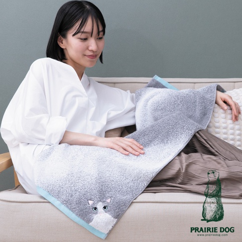 一位年輕女性坐在米色沙發上，身穿白色寬鬆襯衫與棕色長裙，手中輕撫著一條印有可愛貓咪圖案的灰色細柔毛巾。毛巾表面呈現毛絨質感，邊緣配有淺藍色裝飾，右下角帶有 Prairie Dog 品牌圖示與網址。