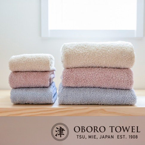 兩疊有機棉浴巾，左疊為米白色、粉色、淺藍色，右疊為米白色、粉色、淺藍色。浴巾質地柔軟，呈現蓬鬆的纖維感。