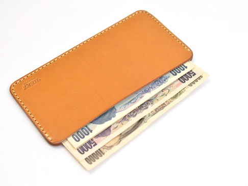 橘色皮革紙鈔收納夾，夾有日幣一千、五千、一萬元紙鈔，夾身有品牌蝕刻標誌，車線工整。