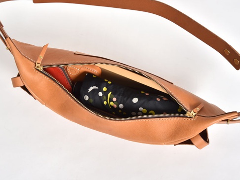 淺棕色皮革大容量斜背包，拉鍊部分敞開，露出內部一個黑色圓點圖案的摺疊傘和一個橘色卡夾。包包配有可調節的同色系皮革肩帶。