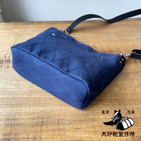 深藍色刺子織迷你側背包，表面有菱格紋縫線，附有黑色織帶和金屬扣環。