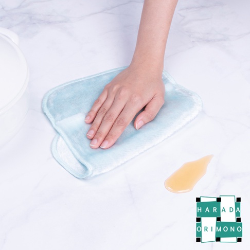 一隻手正用淺藍色的超細纖維布擦拭大理石檯面上的少量黃色液體。