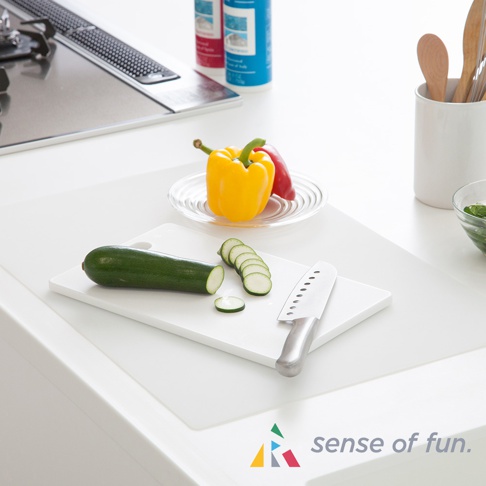 白色耐高溫料理台保護墊，墊上放著一個綠色櫛瓜，部分切片，以及一把銀色刀具。背景有黃色和紅色甜椒，以及廚房爐具。