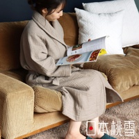 女性身著淺褐色雙面紗布立體織紋浴袍，坐於米色燈芯絨沙發上閱讀雜誌，浴袍材質輕薄，織紋清晰可見，腰間繫有同色系腰帶。